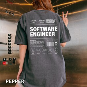 May include: A dark gray t-shirt with the words "SOFTWARE ENGINEER" in large white letters. The shirt has additional white text and graphics, including "100% EXPERTISE" and "INGREDIENTS". The word "PEPPER" is at the bottom of the shirt.