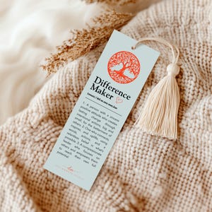 May include: A light blue bookmark with the words "Difference Maker" and a definition. The top features a red tree design and a cream-colored tassel. The bookmark is lying on a cream-colored knitted blanket.