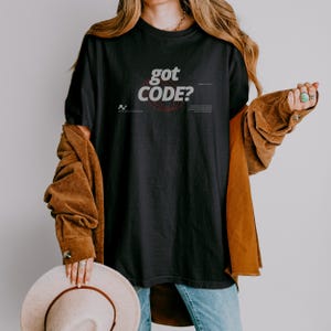 Model wearing a black Comfort Colors 1717 T-shirt with a retro-tech GOT CODE? graphic featuring bold black typography, red orbital linework, and small technical details.