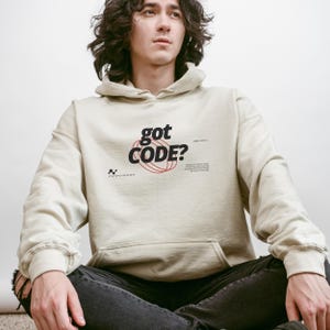 Model wearing a sant colored Gildan G18500 pullover hoodie with a retro-tech graphic design featuring the phrase GOT CODE? in bold typography over red orbital linework with small technical details and humorous token-themed microtext.