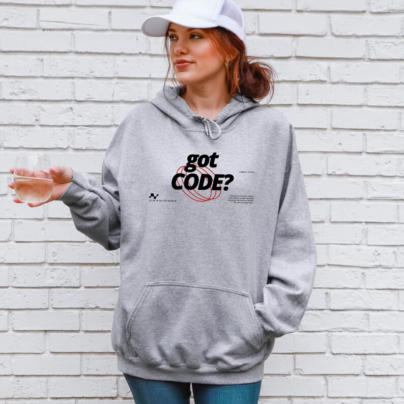 Model wearing a sport grey colored Gildan G18500 pullover hoodie with a retro-tech graphic design featuring the phrase GOT CODE? in bold typography over red orbital linework with small technical details and humorous token-themed microtext.