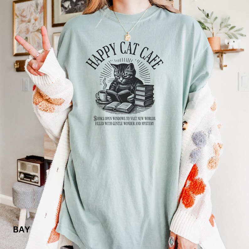 Model wearing oversized bay colored Comfort Colors® T-Shirt with a graphic of a vintage cat reading a book with a cup of coffee and book stack nearby, graphic is styled with Happy Cat Cafe headline and a quirky lost in translation-inspired subhead.