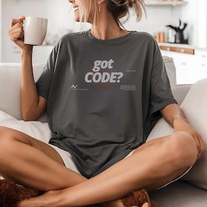 Model wearing a pepper Comfort Colors 1717 T-shirt with a retro-tech GOT CODE? graphic featuring bold black typography, red orbital linework, and small technical details.