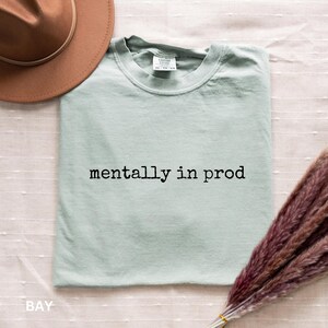 Może przedstawiać: Jasnozielony t-shirt w kolorze szałwiowym z okrągłym dekoltem i czarnym napisem "mentally in prod" na piersi. Koszulka jest pokazana obok brązowego kapelusza z szerokim rondem oraz wiązki suszonej fioletowej trawy pampasowej na białym materiale.