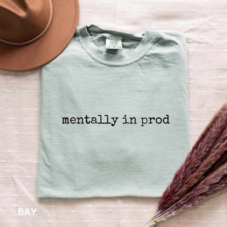 Może przedstawiać: Jasnozielony t-shirt w kolorze szałwiowym z okrągłym dekoltem i czarnym napisem "mentally in prod" na piersi. Koszulka jest pokazana obok brązowego kapelusza z szerokim rondem oraz wiązki suszonej fioletowej trawy pampasowej na białym materiale.