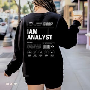 以下が含まれることがあります： 背面にIAM ANALYSTという白いテキストがプリントされた黒の長袖スウェットシャツ。栄養成分表示ラベル風のグラフィックには、様々なアイコン、バーコード、分析職に関連する技術的な説明文が記載されています。