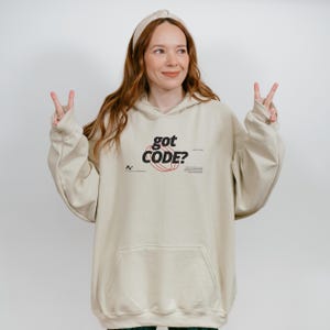Model wearing a sand colored Gildan G18500 pullover hoodie with a retro-tech graphic design featuring the phrase GOT CODE? in bold typography over red orbital linework with small technical details and humorous token-themed microtext.