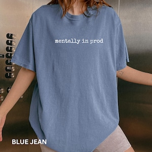 Może przedstawiać: Kr&oacute;tkorękawkowy t-shirt w kolorze dżinsowego błękitu z białym napisem "mentally in prod" nadrukowanym na środku klatki piersiowej. Odzież posiada okrągły dekolt i luźny kr&oacute;j, wykonana z jednolitej, stonowanej niebieskiej tkaniny.