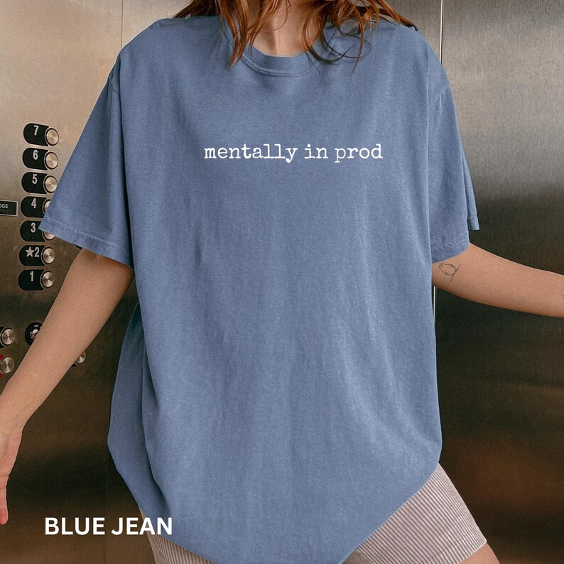 Może przedstawiać: Kr&oacute;tkorękawkowy t-shirt w kolorze dżinsowego błękitu z białym napisem "mentally in prod" nadrukowanym na środku klatki piersiowej. Odzież posiada okrągły dekolt i luźny kr&oacute;j, wykonana z jednolitej, stonowanej niebieskiej tkaniny.