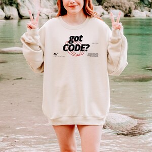 Model wearing a sand colored Gildan G18000 crewneck sweatshirt with a retro-tech graphic design featuring the phrase GOT CODE? in bold typography over red orbital linework with small technical details and humorous token-themed microtext.