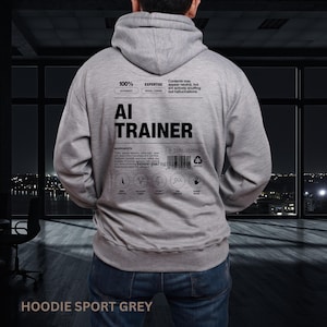 以下が含まれることがあります： 背中に「AI TRAINER」の文字が入ったグレーのパーカー。パーカーにはフードとカンガルーポケットが付いています。「100%」、「EXPERTISE」、「HOODIE SPORT GREY」などのテキストも含まれています。デザインには、材料のリストとアイコンが使用されています。