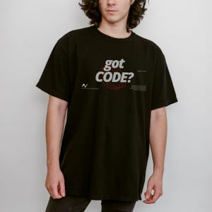 Model wearing a black Comfort Colors 1717 T-shirt with a retro-tech GOT CODE? graphic featuring bold black typography, red orbital linework, and small technical details.