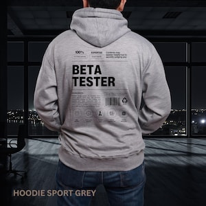 May include: A gray hoodie with the text "BETA TESTER" printed on the back. Additional text includes "100%", "EXPERTISE", and "Contents may appear helpful but is secretly judging you." The hoodie also features a barcode and care symbols. The bottom of the hoodie reads "HOODIE SPORT GREY."