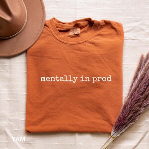 Może przedstawiać: Kr&oacute;tkorękawkowy t-shirt w kolorze wypalonej pomarańczy z białym napisem "mentally in prod" nadrukowanym na klatce piersiowej. Koszulka ma okrągły dekolt i widoczną metkę marki wewnątrz kołnierza. Obok znajduje się brązowy kapelusz i łodyga suszonej rośliny.