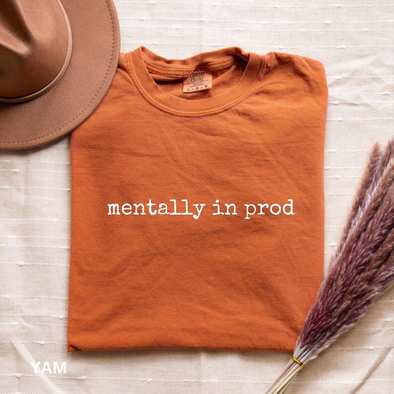 Może przedstawiać: Kr&oacute;tkorękawkowy t-shirt w kolorze wypalonej pomarańczy z białym napisem "mentally in prod" nadrukowanym na klatce piersiowej. Koszulka ma okrągły dekolt i widoczną metkę marki wewnątrz kołnierza. Obok znajduje się brązowy kapelusz i łodyga suszonej rośliny.