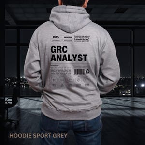 May include: A light gray hooded sweatshirt featuring a black graphic on the back. The design mimics a product label with the text GRC ANALYST, 100% EXPERTISE, and various technical icons. The print includes a barcode and detailed ingredient list.