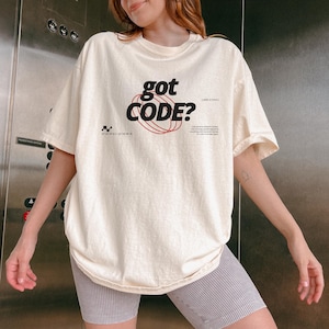 Model wearing an ivory Comfort Colors 1717 T-shirt with a retro-tech GOT CODE? graphic featuring bold black typography, red orbital linework, and small technical details.