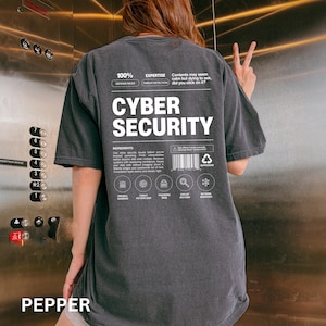 May include: Dark gray t-shirt with the words "CYBER SECURITY" in large white letters. The shirt has additional text and graphics, including "100% DEFENSE MODE" and icons related to cybersecurity. The word "PEPPER" is at the bottom.