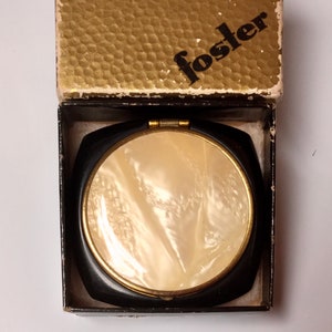 May include: A black compact mirror with a gold trim and a white mother-of-pearl interior. The mirror is in its original box, which is gold with the word "foster" printed in black.