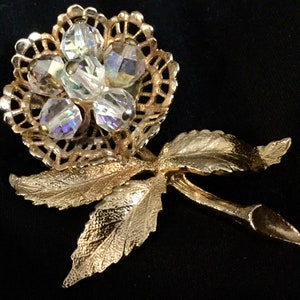 May include: A gold-tone brooch in the shape of a rose with clear iridescent crystals. The rose has a delicate filigree design and is accented with leaves.