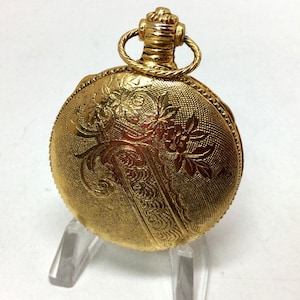 May include: A gold-toned pocket watch with an ornate floral design on the front. The watch has a hinged lid and a decorative chain.