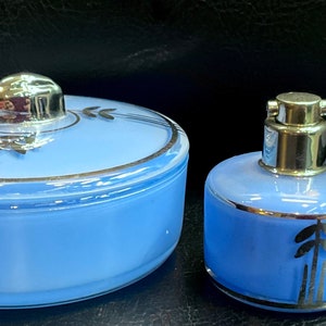 Pode incluir: Um conjunto de toucador vintage azul pó, incluindo um frasco redondo com tampa prateada e um frasco de perfume com atomizador prateado. Ambas as peças têm acabamentos prateados e um design de folha estilizado.
