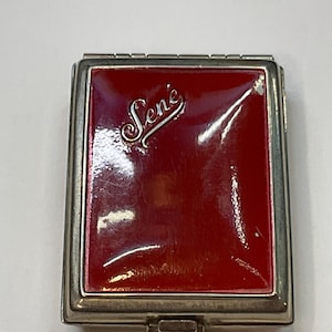 May include: A silver compact with a red enamel top. The compact has the word "Sené" embossed in silver on the top.