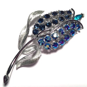 May include: A silver tone brooch with a blue rhinestone flower design. The flower has a long stem with leaves.