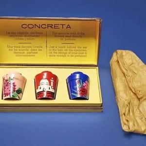 Può includere: Una scatola rettangolare con interno giallo contenente tre piccoli contenitori colorati. Il contenitore a sinistra è rosa con motivi floreali, quello centrale è rosso con il grafico di un edificio bianco e quello a destra è blu con motivi floreali. Un foglio di carta beige stropicciato si trova accanto alla scatola.