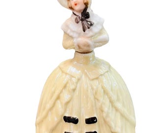 Może przedstawiać: Vintage porcelanowa figurka kobiety w żółtej sukni i kapeluszu. Figurka ma szczegółową twarz, czarną kokardę i czarne akcenty na sukni. Kapelusz ma różowy akcent.