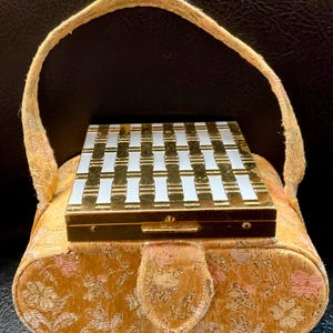May include: A vintage gold and cream-colored handbag with a matching compact. The handbag has a floral pattern and a handle. The compact has a gold frame with a white and gold grid design.