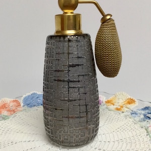 May include: A vintage perfume bottle with a textured, gray glass body and a gold-colored metal top and sprayer. The bottle has a bulb-shaped atomizer. The design is reminiscent of Art Deco style. The bottle is displayed on a white lace doily.