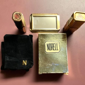 May include: A gold and black vintage perfume set with a small mirror, a black velvet pouch with a gold N, and a bottle of Norell perfume. The perfume bottle has a gold cap and a gold label with the word "Norell" printed in black.
