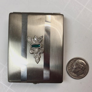 May include: A silver compact with a decorative design featuring a green and clear rhinestone flower. The compact is slightly larger than a US dime.