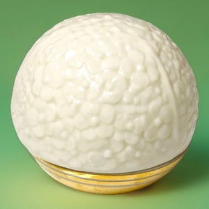Può includere: Oggetto decorativo rotondo, bianco sporco, con superficie strutturata, simile a una pallina da golf. Poggia su una base dorata con tre sottili linee parallele. Lo sfondo è verde unito.
