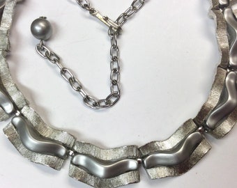 Puede incluir: Un collar de tono plateado con un diseño ondulado. El collar tiene una cadena de tono plateado con un cierre.
