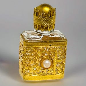 May include: An antique perfume bottle with a gold filigree design. The bottle has a square shape with a clear glass body and a decorative gold cap. A pearl-like embellishment is centered on the front.