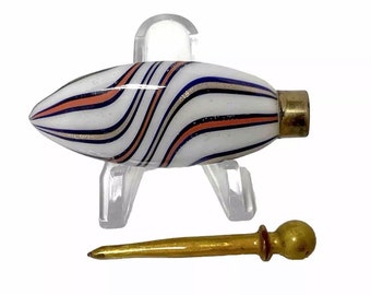 Könnte beinhalten: Eine weiße Glasflasche mit einem wirbelnden Design in Blau, Gold und Rosa. Die Flasche hat einen goldenen Metallverschluss und wird mit einem goldenen Metallapplikator geliefert.
