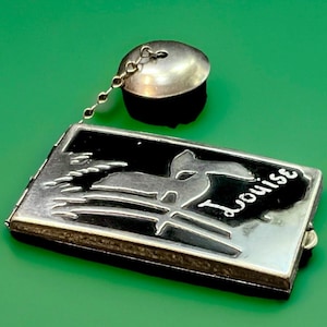 May include: A silver-toned compact with a black background and a stylized design, connected by a chain to a silver-toned lid. The name "Louise" is written in cursive on the compact.