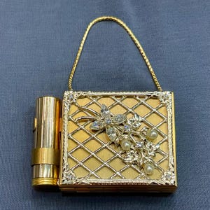 Op de afbeelding: Een vintage goudkleurig compact met een bijpassende lippenstiftkoker. Het compact heeft een traliewerkontwerp met een bloemen- en strass-versiering. Een gouden kettinghandvat is aan het compact bevestigd.