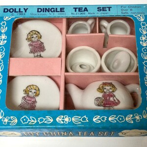 Pode incluir: Um conjunto de chá de brinquedo Dolly Dingle vintage em sua caixa original. O conjunto inclui uma chaleira, xícaras, pires e uma açucareira, todos decorados com uma menina de desenho animado em um vestido rosa. A caixa é azul com detalhes florais e divisórias cor-de-rosa.