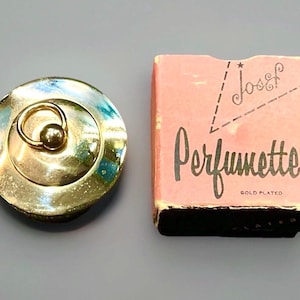 Puede incluir: Un tapón de botella de perfume Perfumette chapado en oro con un asa de anillo y una pequeña bola redonda. Junto a él, una caja rosa con el nombre "Perfumette" en escritura negra y las palabras "Gold Plated".