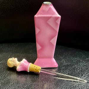 May include: A pink perfume bottle with a geometric design and a silver cap. The bottle is paired with a decorative stopper featuring a small figurine in a pink dress. The stopper has a cork base and a glass applicator.