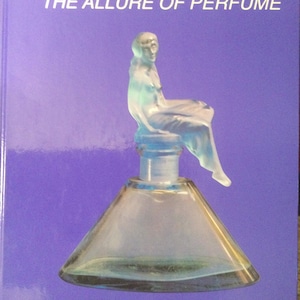 Pode incluir: Um frasco de perfume transparente com uma figura sentada no topo, contra um fundo roxo. O frasco tem uma base cónica. O texto diz "MONSEN AND BAER, Inc. THE ALLURE OF PERFUME" e "Perfume Bottle Auction XVI Washington, D.C. - April 28, 2006."