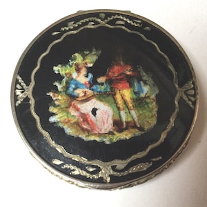 May include: A round, vintage compact with a black enamel surface. The compact features a painted scene of two figures in colorful attire, set against a green and yellow background. The compact is trimmed with a silver-colored metal border and decorative accents.