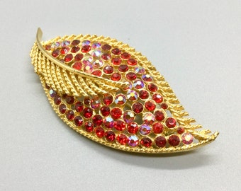 Pode incluir: Broche dourado em forma de folha, adornado com numerosos strass vermelhos e iridescentes. Os strass criam um efeito brilhante. As bordas da folha têm um design texturizado em forma de corda, adicionando detalhes a esta joia vintage.