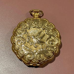Könnte beinhalten: Ein goldfarbener Anhänger mit einem filigranen Blumendesign und einem Vogel im Flug. Der Anhänger hat eine Scharnieröffnung.