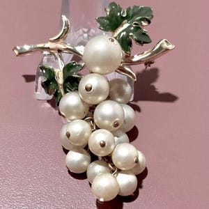 Puede incluir: Un broche de tono dorado con un racimo de perlas falsas, que se asemeja a un racimo de uvas, con hojas de esmalte verde.