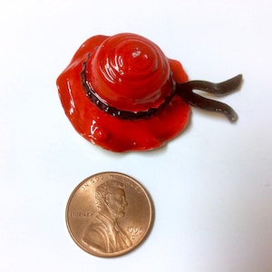 Może przedstawiać: Miniaturowy czerwony i czarny kapelusz z szerokim rondem i czarną wstążką. Kapelusz wykonany jest z błyszczącego materiału i ma błyszczące wykończenie.