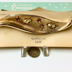 May include: A gold-toned leaf-shaped brooch with textured detailing and sparkling green and amber-colored rhinestones. The brooch is displayed on a vintage card with the text "Sparkling Leaf" and the number "1107".
